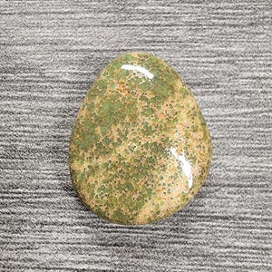 May include: A polished, teardrop-shaped stone with a speckled pattern of green and tan hues. The stone has a glossy finish and a smooth surface, showcasing natural variations in color and texture. The stone is a unique decorative item.