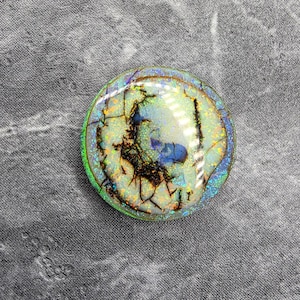 May include: A round, iridescent gemstone with a cracked, web-like pattern in shades of brown, blue, and green. The gemstone is set against a dark gray, textured background. The gemstone has a reflective, polished surface.