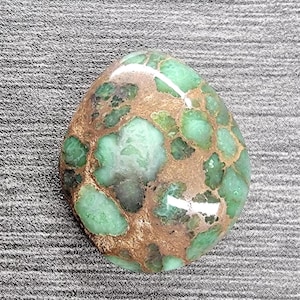May include: A green and brown variscite cabochon with a smooth, polished surface. The stone has a unique pattern of green and brown swirls.