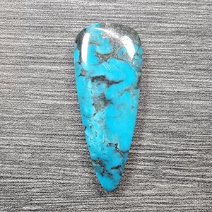 May include: A teardrop-shaped turquoise cabochon with a blue and black marbled pattern. The stone is polished and has a smooth, glossy finish.