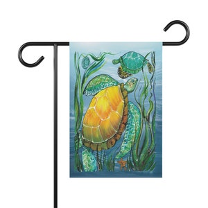 May include: A garden flag depicting an underwater scene with two sea turtles. The turtles have yellow and green shells, amidst green seaweed against a blue backdrop. The flag is mounted on a black metal stand.