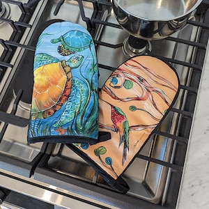 May include: Two colorful oven mitts with a black trim. One mitt features a watercolor painting of a turtle in a blue and green ocean scene. The other mitt features a watercolor painting of a hummingbird on a branch with a tan and orange background. The text "Sharon Davis" is printed on the hummingbird mitt.