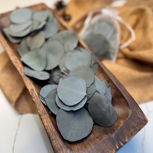 May include: A wooden bowl filled with dried eucalyptus leaves. The leaves are a light gray-green color and are arranged in a pile.