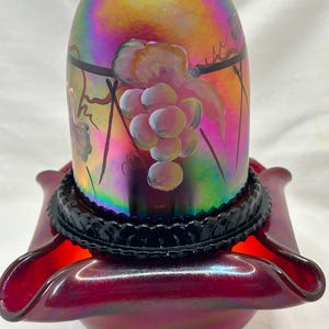 May include: A red iridescent glass lamp shade featuring a hand-painted grape cluster design with green leaves and brown vines. The shade rests in a black scalloped metal holder set atop a deep red ruffled glass base with a metallic sheen.