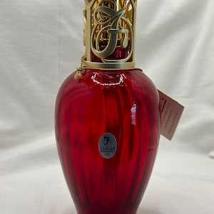 May include: A red glass Fenton fragrance lamp with a gold-colored filigree top. The lamp has a rounded base and a textured, ribbed design. The top features the letter "F". A small tag is attached to the lamp.