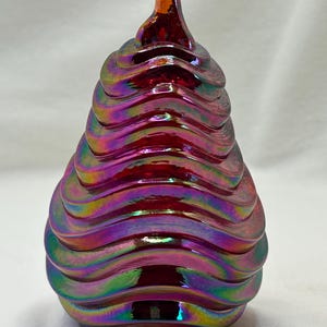 May include: A decorative glass sculpture with a swirling, layered design. The sculpture features iridescent colors of red, purple, and green. The top has a small, curved, amber-colored tip.