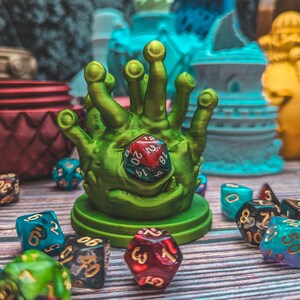 Puede incluir: Una torre de dados de mano de color verde, impresa en 3D, con un dado d20 rojo y verde en su interior. La torre está rodeada de varios dados multicolores.