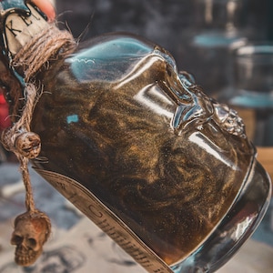 Puede incluir: Una botella de vidrio con un diseño en forma de calavera, llena de un líquido dorado brillante. La botella está atada con una cuerda y tiene un pequeño amuleto de calavera colgando.
