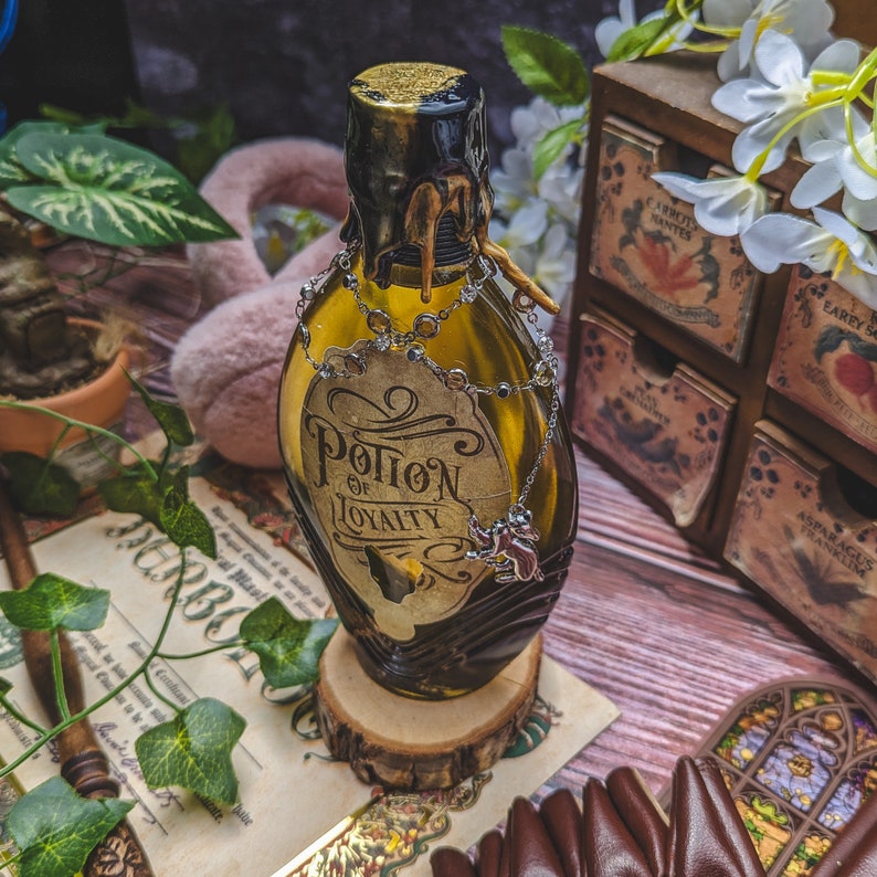 Może przedstawiać: Dekoracyjna, stylizowana na antyczną szklana butelka ze złotą zakrętką i etykietą z napisem "Potion of Loyalty". Butelka ozdobiona jest srebrnym łańcuszkiem i małym charms'em. Stoi na drewnianej podstawie, sugerując motyw vintage lub fantasy.