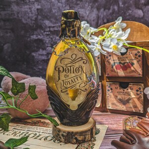 Może przedstawiać: Bursztynowa butelka z eliksirem ze złotą etykietą z napisem "Potion of Loyalty". Butelka ma ciemnobrązową podstawę i złotą g&oacute;rę. Stoi na drewnianym plastrze.