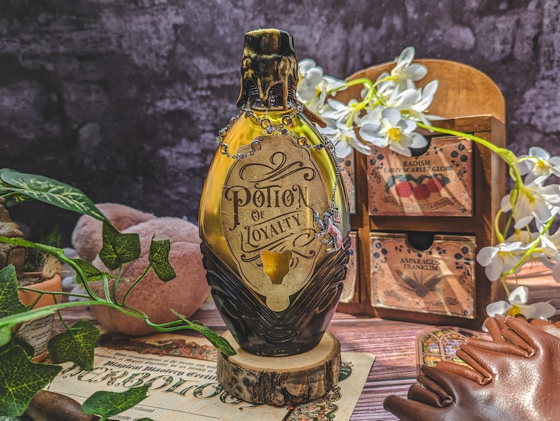 Może przedstawiać: Bursztynowa butelka z eliksirem ze złotą etykietą z napisem "Potion of Loyalty". Butelka ma ciemnobrązową podstawę i złotą g&oacute;rę. Stoi na drewnianym plastrze.