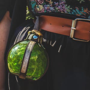 Könnte beinhalten: Eine grüne Glas-Trankflasche mit einem dekorativen goldfarbenen Deckel und einem Lederriemen. Die Flasche ist mit "GREENWELD POTION" beschriftet und an einem braunen Ledergürtel mit einer goldenen Schnalle befestigt. Das Outfit ist schwarz.