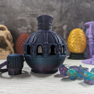 Op de afbeelding: Een donkerblauwe en paarse iriserende fles met een gedetailleerd ontwerp en een bijpassende dop. De fles wordt omringd door verschillende blauwe en paarse dobbelstenen met gouden cijfers.