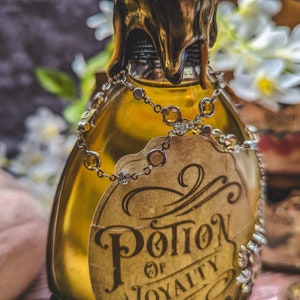 Może przedstawiać: Ozdobna szklana butelka wypełniona złotym płynem, ozdobiona etykietą z napisem "Potion of Loyalty". Butelka jest ozdobiona łańcuszkiem z biżuterią i złotą pieczęcią woskową. W tle białe kwiaty i drewniane pudełko.