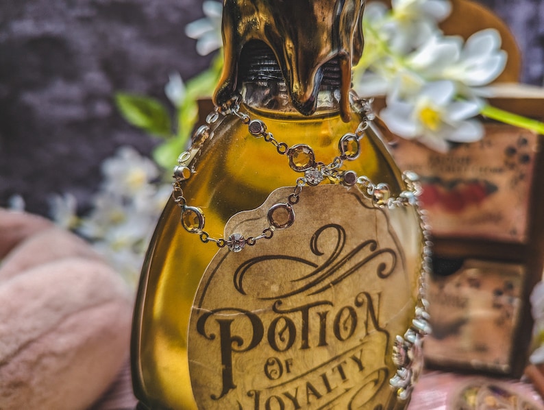 Może przedstawiać: Ozdobna szklana butelka wypełniona złotym płynem, ozdobiona etykietą z napisem "Potion of Loyalty". Butelka jest ozdobiona łańcuszkiem z biżuterią i złotą pieczęcią woskową. W tle białe kwiaty i drewniane pudełko.