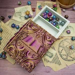 May include: A white wooden box with a hinged lid, decorated with gold and purple floral patterns. The box is open, revealing a compartment filled with colourful dice. The box is sitting on a wooden surface with old paper and dice scattered around it.