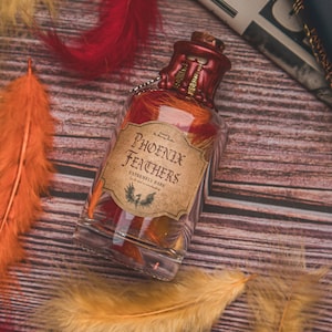 Könnte beinhalten: Eine Glasflasche gefüllt mit roten und orangefarbenen Federn, mit einem Etikett mit der Aufschrift "Phoenix Feathers Extremely Rare". Die Flasche steht auf einer Holzoberfläche mit orangefarbenen, roten und gelben Federn, die um sie herum verstreut sind.