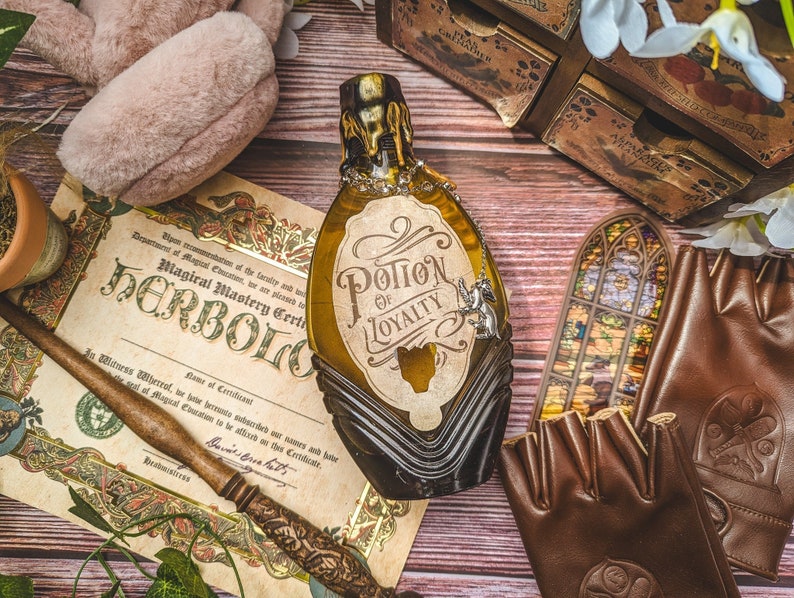 Może przedstawiać: Butelka z miksturą w stylu antycznym z napisem "Potion of Loyalty" z ozdobnym łańcuszkiem i zawieszką. Obraz zawiera certyfikat, r&oacute;żdżkę, brązowe rękawiczki bez palc&oacute;w i małe drewniane pudełko, wszystko ułożone na drewnianej powierzchni.