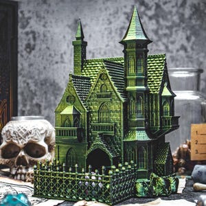 May include: A green 3D printed miniature haunted house with a fence and a detailed exterior. The house has a pointed roof, multiple windows, and a small door. The house is sitting on a table with other miniature items.