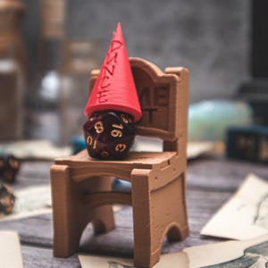 Könnte beinhalten: Ein kleiner brauner Holzstuhl trägt einen dunkelroten, vielseitigen Würfel mit weißen Zahlen. Ein roter kegelförmiger Hut mit dem Wort "AZURE" befindet sich auf dem Würfel. Die Szene spielt sich auf einer Holzoberfläche mit anderen Objekten im Hintergrund ab.