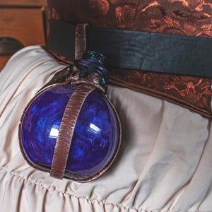Könnte beinhalten: Eine runde Glasflasche mit einer tiefblauen, wirbelnden Flüssigkeit im Inneren, gehalten von einem braunen Lederriemen und Metallakzenten. Die Flasche ist Teil eines Kostüms, möglicherweise für ein Fantasy- oder Steampunk-Thema.