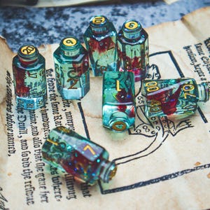 Può includere: Piccole bottiglie di vetro trasparente con tappi dorati e numeri, riempite con una sostanza rossa e dorata. Sono disposte su carta antica con testo, creando un'estetica vintage.