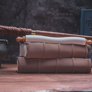 May include: A wooden wand sits atop a stack of three brown leather-bound books. A white scroll is wrapped around the books. The top book has the title "Perfect Potions" on its cover.