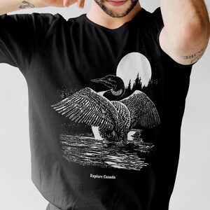 May include: Black t-shirt featuring a white graphic of a loon with its wings spread, standing in water with a full moon and trees in the background. The text "Explore Canada" is printed below the loon.