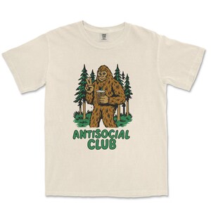 May include: Cream-colored t-shirt featuring a cartoon Bigfoot character holding a beverage and making a peace sign. The Bigfoot is brown with green grass and pine trees in the background. The words "ANTISOCIAL CLUB" are printed in green below the graphic.