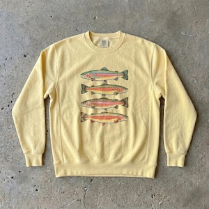 Peut inclure: Un sweat-shirt à col rond et manches longues de couleur jaune pâle, orné d'une impression graphique représentant quatre truites arc-en-ciel empilées. Les poissons sont illustrés dans des tons de vert, rose et orange. Col, poignets et ourlet côtelés.