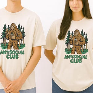 May include: Two cream-colored t-shirts with a Bigfoot graphic. The Bigfoot is brown and holding a beverage, giving a peace sign. Behind the Bigfoot are green trees. The text "ANTISOCIAL CLUB" is in green below the graphic.