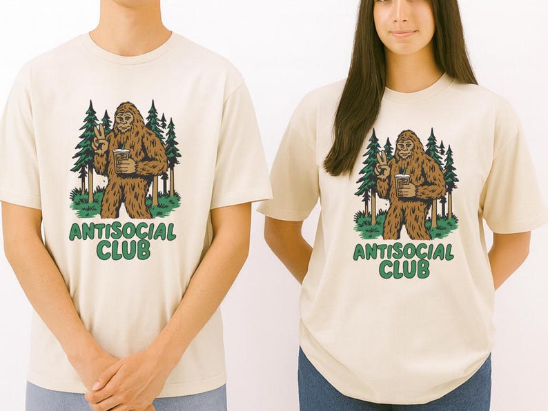 May include: Two cream-colored t-shirts with a Bigfoot graphic. The Bigfoot is brown and holding a beverage, giving a peace sign. Behind the Bigfoot are green trees. The text "ANTISOCIAL CLUB" is in green below the graphic.
