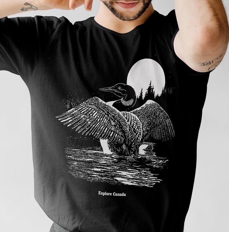 May include: Black t-shirt featuring a white graphic of a loon with its wings spread, standing in water with a full moon and trees in the background. The text "Explore Canada" is printed below the loon.
