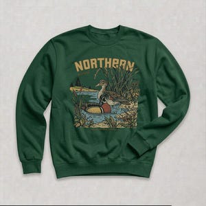 May include: A dark green crewneck sweatshirt featuring a graphic illustration of two ducks swimming in a pond surrounded by reeds. The text NORTHERN is printed in a tan, arched font above the ducks, with the word marsh written in smaller script.