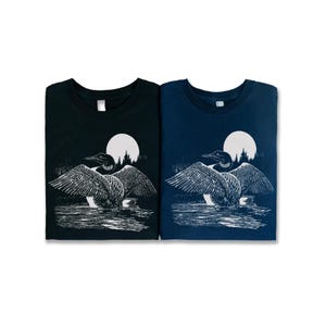 May include: Two t-shirts featuring a white graphic of a loon with its wings outstretched, standing in water with a full moon and trees in the background. The t-shirt on the left is black and the t-shirt on the right is navy blue.