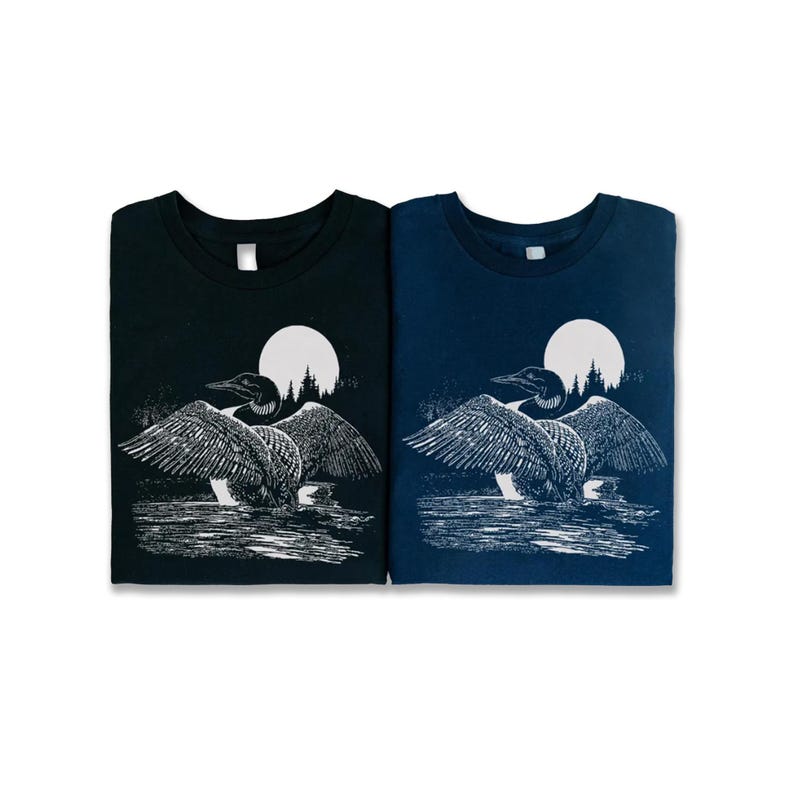 May include: Two t-shirts featuring a white graphic of a loon with its wings outstretched, standing in water with a full moon and trees in the background. The t-shirt on the left is black and the t-shirt on the right is navy blue.