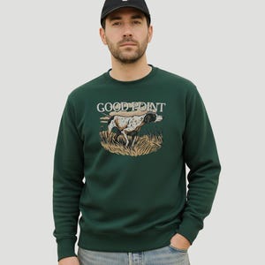 Peut inclure: Un sweat-shirt vert foncé avec l'inscription "GOOD POINT" et une illustration d'un chien de chasse. La personne porte une casquette de baseball noire avec un "C" blanc et un jean bleu.