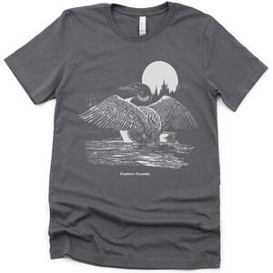 May include: A dark grey t-shirt featuring a white graphic of a loon in front of a full moon and trees. The text "Explore Canada" is printed below the loon.
