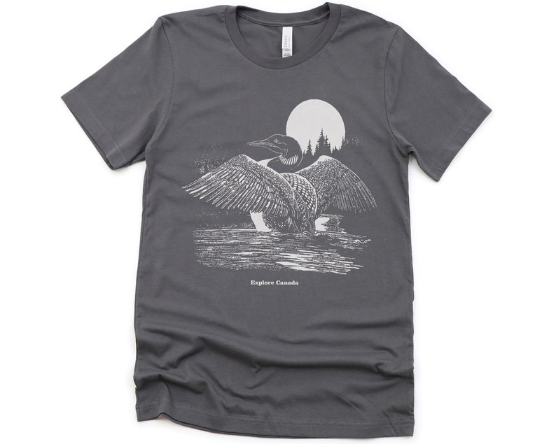 May include: A dark grey t-shirt featuring a white graphic of a loon in front of a full moon and trees. The text "Explore Canada" is printed below the loon.