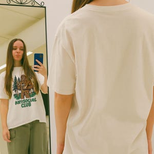 May include: Cream-colored t-shirt with a graphic of a Bigfoot-like creature holding a cup, with trees and the text "ANTISOCIAL CLUB" in green. The shirt is worn by a person in front of a mirror.