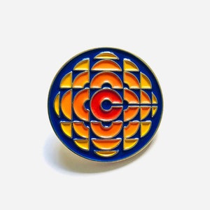 Pode incluir: Um broche de esmalte azul e laranja com o icônico logotipo da CBC, um "C" estilizado cercado por círculos concêntricos.