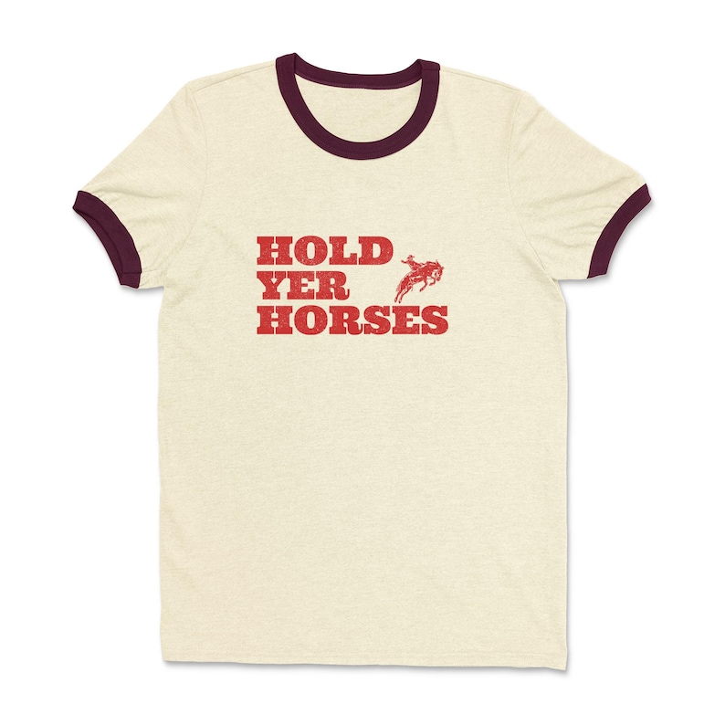 以下が含まれることがあります： マロン色のリブ付きのベージュのTシャツ。シャツには「HOLD YER HORSES」という赤いグラフィックと、馬に乗ったカウボーイの小さな画像が描かれています。