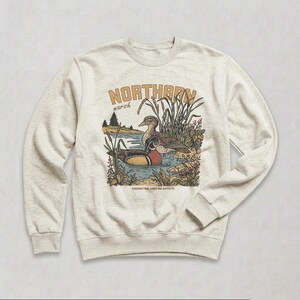 May include: A heathered oatmeal crewneck sweatshirt featuring a graphic illustration of two ducks in a marsh. The text reads NORTHERN marsh and CANADIAN TAWN GOODS AND SUPPLY CO. The design includes cattails, reeds, and pine trees in muted tones.