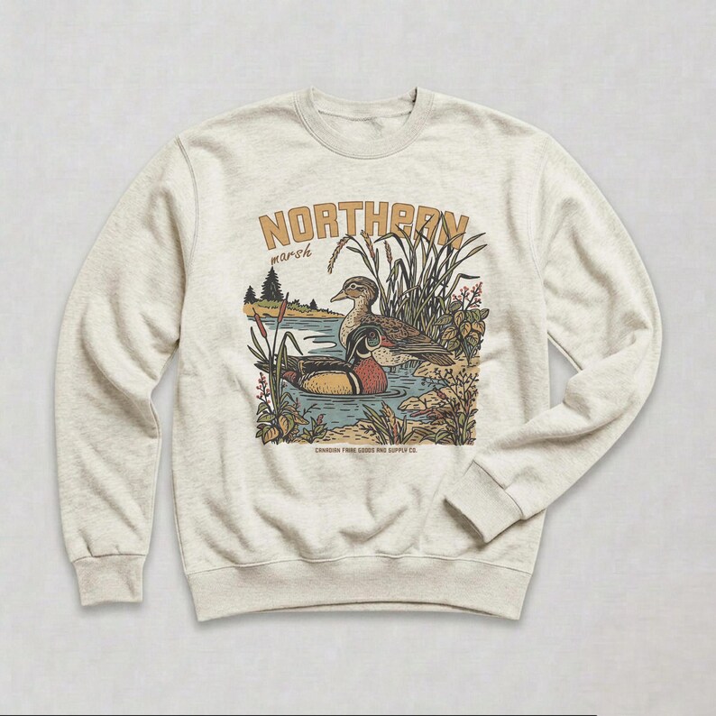 May include: A heathered oatmeal crewneck sweatshirt featuring a graphic illustration of two ducks in a marsh. The text reads NORTHERN marsh and CANADIAN TAWN GOODS AND SUPPLY CO. The design includes cattails, reeds, and pine trees in muted tones.