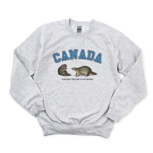 May include: A light gray crewneck sweatshirt featuring the word CANADA in blue block letters with a black outline. Below the text are two illustrated raccoons facing each other, with the phrase EXPLORE THE GREAT OUTDOORS printed in black.