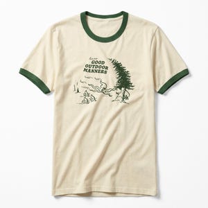 May include: Cream-coloured t-shirt with a green collar and sleeve trim. The shirt features a green graphic of a tree, river, and text that reads "have good outdoor manners".