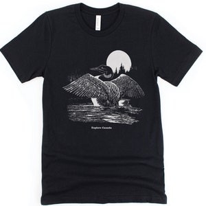 May include: Black t-shirt featuring a white graphic of a loon in front of a full moon and trees. The text "Explore Canada" is printed below the loon.