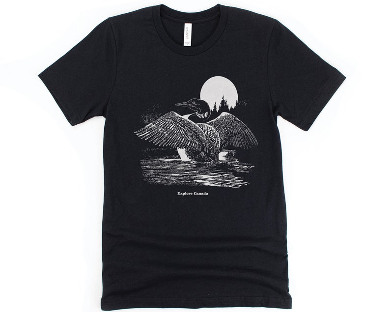 May include: Black t-shirt featuring a white graphic of a loon in front of a full moon and trees. The text "Explore Canada" is printed below the loon.