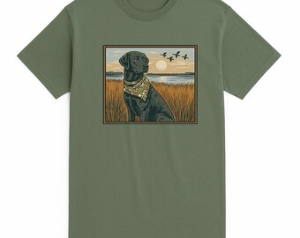 May include: Olive green t-shirt featuring a graphic of a black Labrador wearing a camouflage bandana, sitting in tall grass, with a sunset and flying birds in the background. The shirt is made of a soft material.