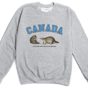 May include: A heather gray crewneck sweatshirt featuring the word CANADA in blue block letters with black outlines. Below the text are two raccoons and the phrase EXPLORE THE GREAT OUTDOORS printed in small black capital letters on the chest.